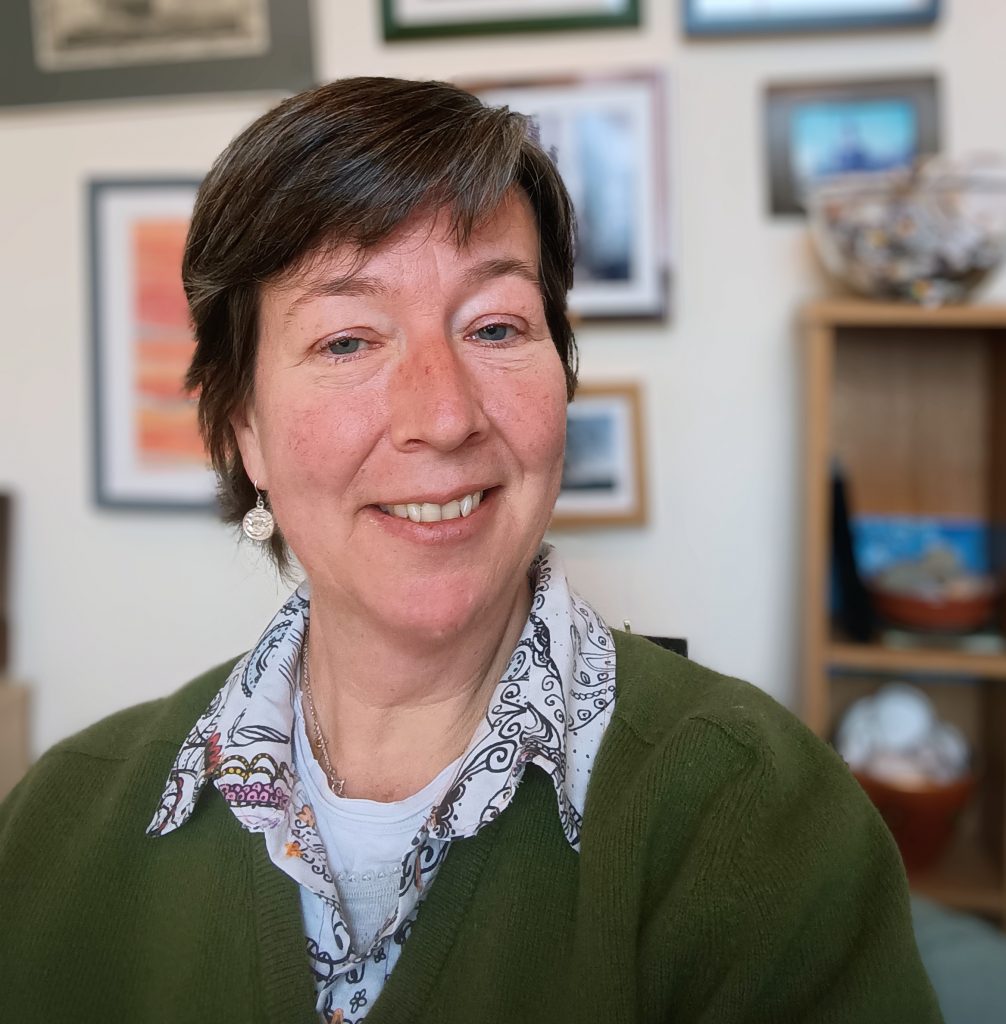 Photo of me, a white middle aged woman with short brown hair. I am smiling, sitting in front of a wall of slightly crooked artwork.