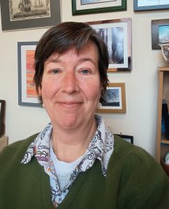 Photo of me, a white middle aged woman with short brown hair. I am smiling, sitting in front of a wall of slightly crooked artwork.