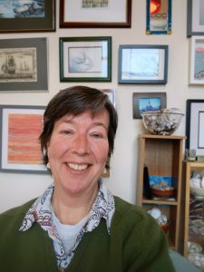 Photo of me, a white middle aged woman with short brown hair. I am smiling, sitting in front of a wall of slightly crooked artwork.