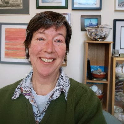 Photo of me, a white middle aged woman with short brown hair. I am smiling, sitting in front of a wall of slightly crooked artwork.